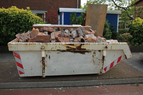 A white metal skip container filled with a large pile of broken red bricks and rubble, positioned on a paved residential driveway near a garden hedge and a blue shed. The skip is weathered with visible rust and stains, and is situated in an outdoor setting in Hendon, London. Surrounding greenery includes shrubs and trees, indicating a suburban environment. The scene relates to waste removal services provided by Commercial Waste Hendon, illustrating the collection of construction debris or renovation waste typical for rubbish clearance in the area.