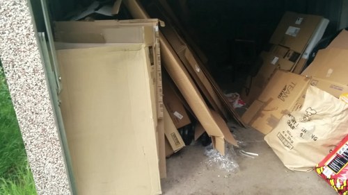 A storage area in an outdoor or semi-enclosed space showing a collection of various waste materials and discarded items. In the foreground on the left, there is a beige shelving unit or cabinet with a smooth surface and slightly reflective finish, leaning against a wall with a speckled texture. To the right, there are multiple cardboard boxes in different sizes, some partially crushed, with printed labels and logos, stacked or piled haphazardly. Among the boxes, there are loose pieces of cardboard, plastic wrapping, and protective bubble wrap. The background includes more cardboard boxes and paper waste, some leaning or stacked atop each other, creating a cluttered scene. The ground appears to be concrete or paving, and the setting looks like an outdoor storage area or yard near residential or commercial premises in Hendon, London, with natural light illuminating the scene. This image is relevant to rubbish removal services offered by Commercial Waste Hendon, illustrating typical waste items that may require collection.