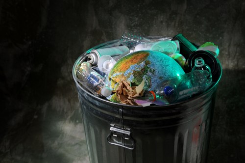 A metal rubbish bin with a secure latch lid, filled with various discarded plastic bottles, containers, and crumpled paper, including a small globe or planet Earth model among the waste. The bin is positioned outdoors on a paved surface, possibly a driveway or pavement in Hendon, with a dark, textured background. The plastic bottles and containers are of different shapes, sizes, and colours, predominantly transparent or light green, reflecting the ambient light. The crumpled paper appears brown and is partially visible among the plastics. This image relates to waste disposal and rubbish collection services, such as those offered by Commercial Waste Hendon, highlighting the importance of proper rubbish management in local areas.